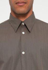 Camisa gris con cuello recto y rayas verticales, con dos botones de cierre; la tela tiene una textura suave.