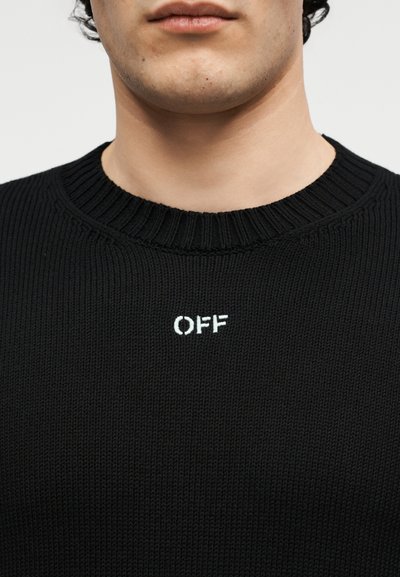 Κοντινό πλάνο ενός ατόμου που φοράει μαύρο πλεκτό πουλόβερ με ριμπ γιακά και λευκό κεντημένο κείμενο «OFF» στο στήθος.