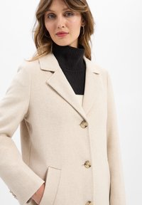 Cappotto in lana beige con un design strutturato, rever a punta e bottoni grandi. Presenta tasche laterali e un dolcevita nero testurizzato sotto.