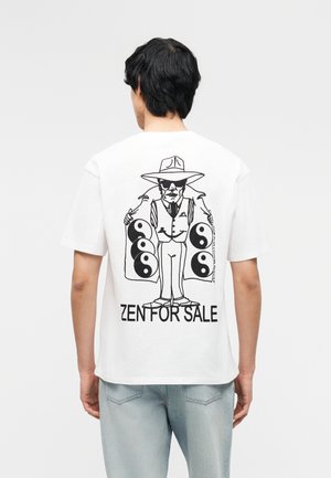 Mężczyzna w białej koszulce z czarną grafiką przedstawiającą mężczyznę w kapeluszu i okularach przeciwsłonecznych trzymającego otwarty płaszcz pokazujący symbole yin-yang, poniżej napis „ZEN FOR SALE”.