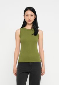 Jonge vrouw met lang zwart haar draagt een mouwloze olijfgroene top en zwarte broek, staand tegen een effen witte achtergrond.