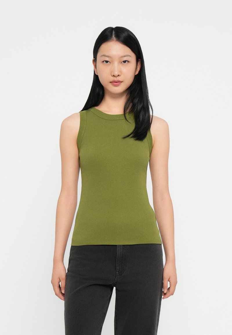 Jonge vrouw met lang zwart haar draagt een mouwloze olijfgroene top en zwarte broek, staand tegen een effen witte achtergrond.