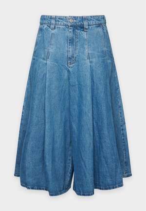 Une femme porte un chemisier crème à nœud au col, une jupe midi en jean bleu, une ceinture marron et des sandales noires sur un fond clair uni.