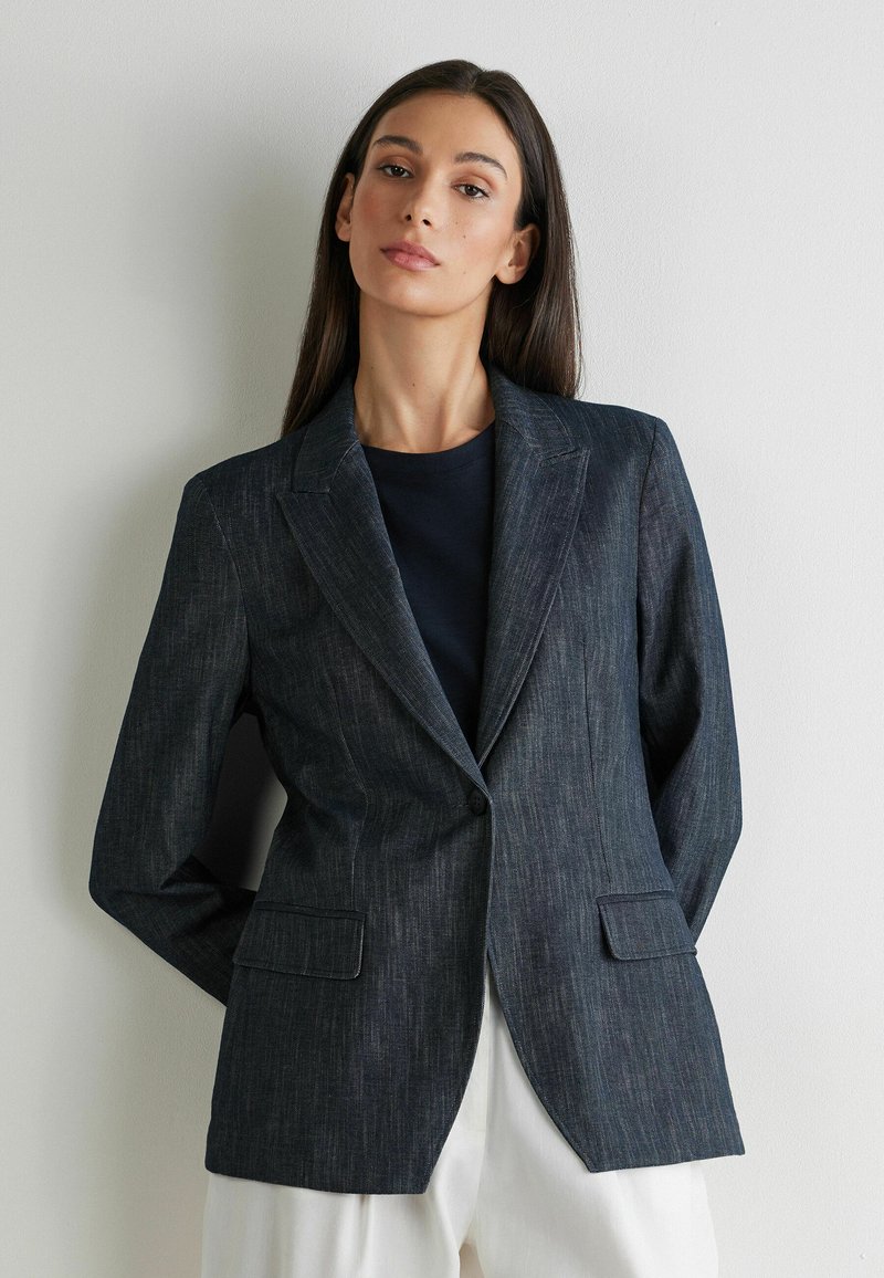 Femme aux longs cheveux foncés portant un blazer à fines rayures sombre sur une chemise noire et un pantalon blanc, debout contre un mur blanc uni.