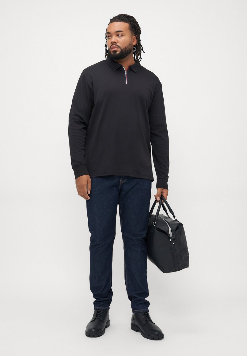 Schwarzes langärmliges Polohemd mit Reißverschlusskragen, kombiniert mit dunkelblauen Jeans und schwarzen Lederstiefeln, hält eine strukturierte schwarze Tasche.