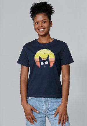 T-shirt en coton bleu marine arborant un graphisme d'un chat noir aux grands yeux, sur un dégradé circulaire de coucher de soleil en jaune et orange.