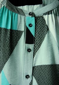 Camicia pulsante in tessuto turchese e nero con motivi geometrici, colletto rotondo e bottoni neri lungo la patta frontale.