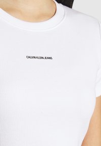 White ribbed t-shirt with a round neck, featuring the text "CALVIN KLEIN JEANS" in black, positioned on the chest area.