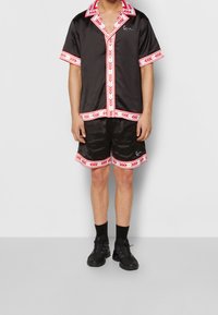 Person wearing black satin short-sleeve shirt and matching shorts with red and white patterned trim, black socks, and black athletic shoes.