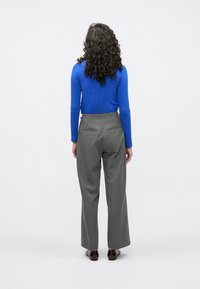 Šedé šité kalhoty a modrý přiléhavý top s dlouhým rukávem, s přímým střihem a hladkou texturou. Pohled zezadu s kudrnatými vlasy.