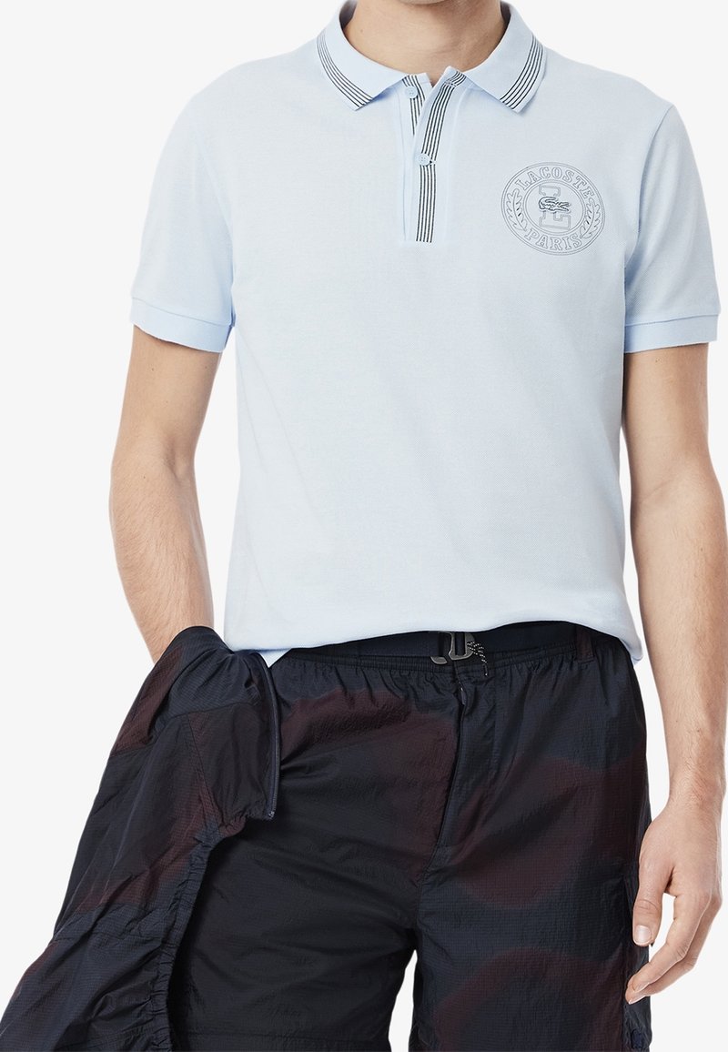 Homme portant un polo Lacoste bleu clair avec col rayé et un short foncé tenant une veste sombre dans une main.