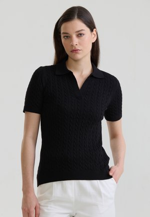 Frau trägt ein schwarzes kurzärmeliges Kabelstrick-Poloshirt mit weißen Hosen, steht mit einer Hand in der Tasche vor einem einfarbigen Hintergrund.
