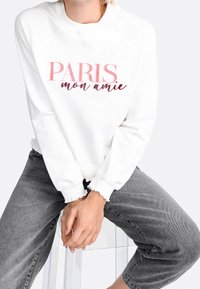 Weißes Sweatshirt mit "PARIS mon amie" in pinker Schrift, mit lockerer Passform, gerippten Bündchen und ausgefranstem Saum, kombiniert mit grauen Jeans.