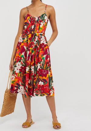 Vrouw draagt een mouwloze midi-jurk met ceintuur, bedrukt met rode, witte, groene en gele bloemen, houdt een gevlochten tas vast en draagt mosterdkleurige slippers.