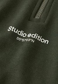 Olivgrüner Reißverschluss-Fleece mit weißem Text "Studio-Edition" und den Koordinaten "55°5703''N" auf der Vorderseite, mit einer weichen Textur.
