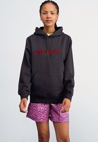Black hoodie with red "Carhartt" logo, front pouch pocket, paired with pink leopard print shorts featuring a small blue tag.