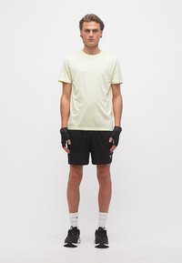 Un jeune homme se tient face à l'avant, portant un t-shirt jaune pâle, un short noir, des gants sans doigts, des chaussettes blanches et des chaussures de sport noires.
