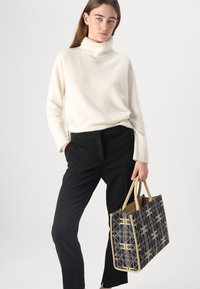 Pullover a collo alto color crema, con polsini a coste, abbinato a pantaloni neri. Tiene in mano una borsa a tracolla a motivi blu navy con rifiniture beige e manici.