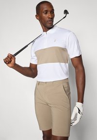 Golfantrekk med en hvit kortermet polo med en beige stripe og beige shorts. Modellen holder en golfkølle og har på seg en hvit golfhanske.