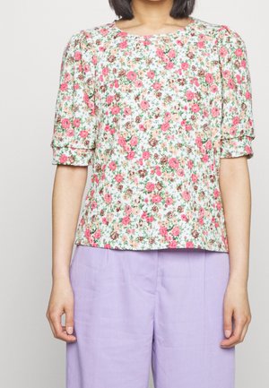 Personne portant un chemisier blanc à manches courtes avec un imprimé floral rose et un pantalon large violet clair, debout devant un fond uni.
