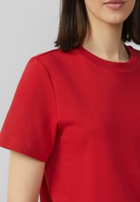 Rode katoenen t-shirt met korte mouwen. Ronde halslijn en schoudernaden van detail. Gladde textuur, geen zichtbare patronen. Close-up weergave.