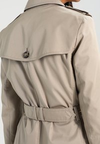 Zbliżenie na osobę noszącą beżowy trencz z tylną yoke, widocznymi detalami guzików i paskiem w talii.