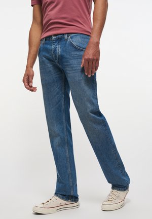 Jeans de denim azul oscuro con un diseño de pierna recta, costuras visibles y áreas desgastadas, combinados con zapatillas de lona de color claro.