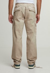Beige cargo-housut rentoa istuvuutta, elastinen vyötärö ja useita taskuja. Kangas näyttää pehmeältä ja hieman teksturoidulta. Kengät näkyvät alareunassa.