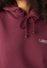 Primer plano de una persona que lleva una sudadera burdeos con cordones ajustados y texto bordado blanco parcial en el pecho.