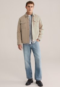 Beige geïsoleerde jas met twee borstzakken, gedragen over een licht geruit shirt, gecombineerd met lichtblauwe jeans en zwarte laarzen.