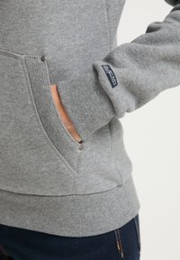 Šedá mikina s vroubkovaným lemem a manžetami, boční kapsou s knoflíkovým akcentem a štítkem na rukávu. Měkká textura.