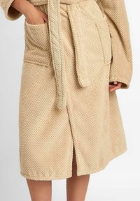 Beżowy szlafrok z dzianiny frotté o teksturowanym splocie, z paskiem w talii, dwoma przednimi kieszeniami, sięgający do kolan i z rozcięciem na dole dla ułatwienia ruchów.