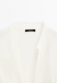 Blouse blanche à la texture douce, avec un col en V et des détails de couture subtils. L'étiquette indique « Massimo Dutti, Fabriqué en Chine. »