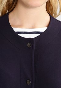 Donkerblauwe cardigan met een ronde halslijn, voorzien van een enkele zwarte knoopsluiting. Daaronder is een wit shirt met donkerblauwe strepen zichtbaar.