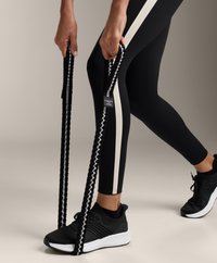 Crne otporne trake držane u rukama, s teksturiranim hvataljkama. Crne tajice s kremastim prugama i crne sportske cipele s bijelim đonovima.