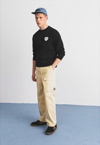 Svart langermet skjorte med hjerte-logo, beige cargo bukser med lommer, og svarte joggesko med brune såler. Brukt med en lyseblå caps.