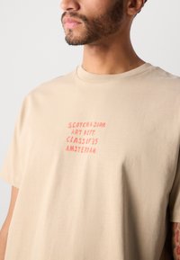 Beige pamut póló piros felirattal: "SCOTCH & SODA ART DEPT. CLASS OF '85 AMSTERDAM." Kerek nyakú, rövid ujjú, sima textúrájú.