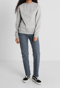 Mujer vestida con un sudadera gris claro, jeans azules y zapatillas negras y blancas, de pie contra un fondo liso claro.