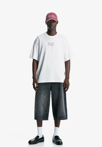 Valkoinen oversized T-paita, jossa on painettua tekstiä, yhdistettynä tummiin denim-culotteihin. Mustat loaferit ja valkoiset sukat täydentävät asua, johon kuuluu myös vaaleanpunainen lippis.