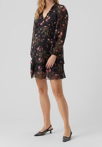 Mustakukkainen mekko, jossa on pitkät läpinäkyvät hihat, kerroksellinen hame ja V-pääntie. Mekko sisältää vaaleanpunaisia ja vihreitä kukkakuosia tummalla taustalla.