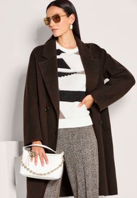 Frau trägt einen oversized, dunkelbraunen Mantel, einen gemusterten Pullover, karierte Hosen, getönte Sonnenbrillen und hält eine weiße geflochtene Handtasche mit goldener Kette.