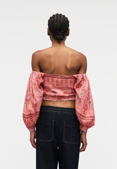Femme aux cheveux tressés coiffés en chignon, portant un chemisier rouge à motifs, épaules nues, manches bouffantes et un pantalon large sombre, vue de dos.