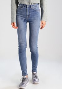 Jeans ajustados de cintura alta en azul claro con un lavado desgastado, que presentan un cierre frontal de botón y cinco bolsillos, combinados con zapatillas grises.