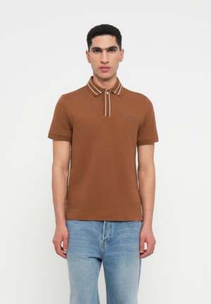 Polo shirt marron avec un col côtelé présentant des rayures blanches et un petit logo tissé sur la poitrine. Porté avec un jean bleu clair.