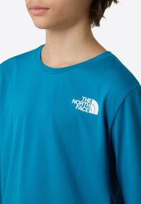 Svetlo modra majica s kratkimi rokavi in okroglim izrezom, izdelana iz teksturirane tkanine, z belim logotipom na levi strani prsnega koša. Kratki rokavi in sproščen kroj.