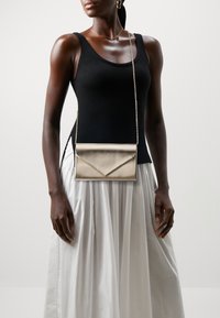 Žena v černém bezrámovém topu a bílé sukni, doplněná zlatou kabelkou a zlatými náušnicemi na jednolitém pozadí.