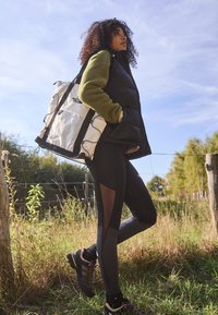 Fekete pufi mellény zöld fleece fölött, fekete leggings hálós panelekkel, szürke túracipő és egy fehér hátizsák fekete részletekkel.