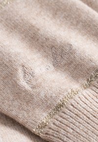 Beige gebreide trui met een zachte textuur, subtiele geribbelde zoom en decoratieve gouden draadaccenten. Borduursel met logo aanwezig.
