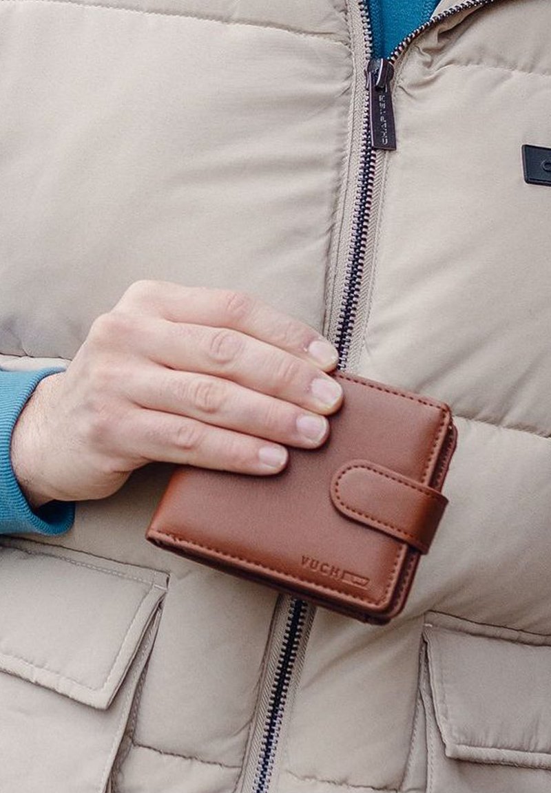 Brown leather wallet with a clasp closure, held in a hand. Features a smooth texture, rectangular shape, and embossed brand name.