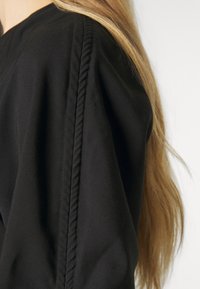 Gros plan d'une personne aux longs cheveux blonds portant un vêtement noir avec un détail de couture verticale texturée sur la manche.
